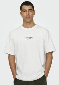 Weißes Baumwoll-T-Shirt mit kurzen Ärmeln und Rundhalsausschnitt, mit schwarzem Text "CURATED STAPLES" im Brustbereich.