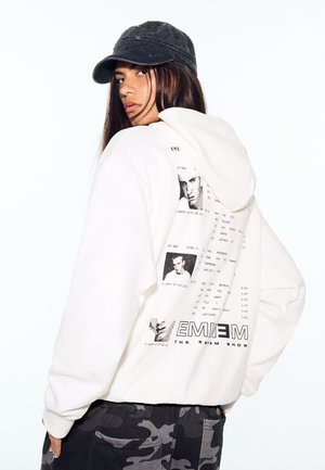 Jeune personne portant un sweat à capuche blanc avec la pochette d'album d'Eminem dans le dos et une casquette noire, regardant par-dessus son épaule sur un fond blanc.