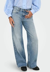 Vaqueros de pierna ancha en azul claro, fabricados en denim, con cierre de botón, cinco bolsillos y un lavado desgastado, combinados con una camisa de corte holgado.