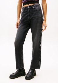 Jeans de mezclilla negros de tiro alto con corte recto, que presentan un detalle de tachuelas a lo largo del costado y una textura sutil. Combinados con mocasines negros.