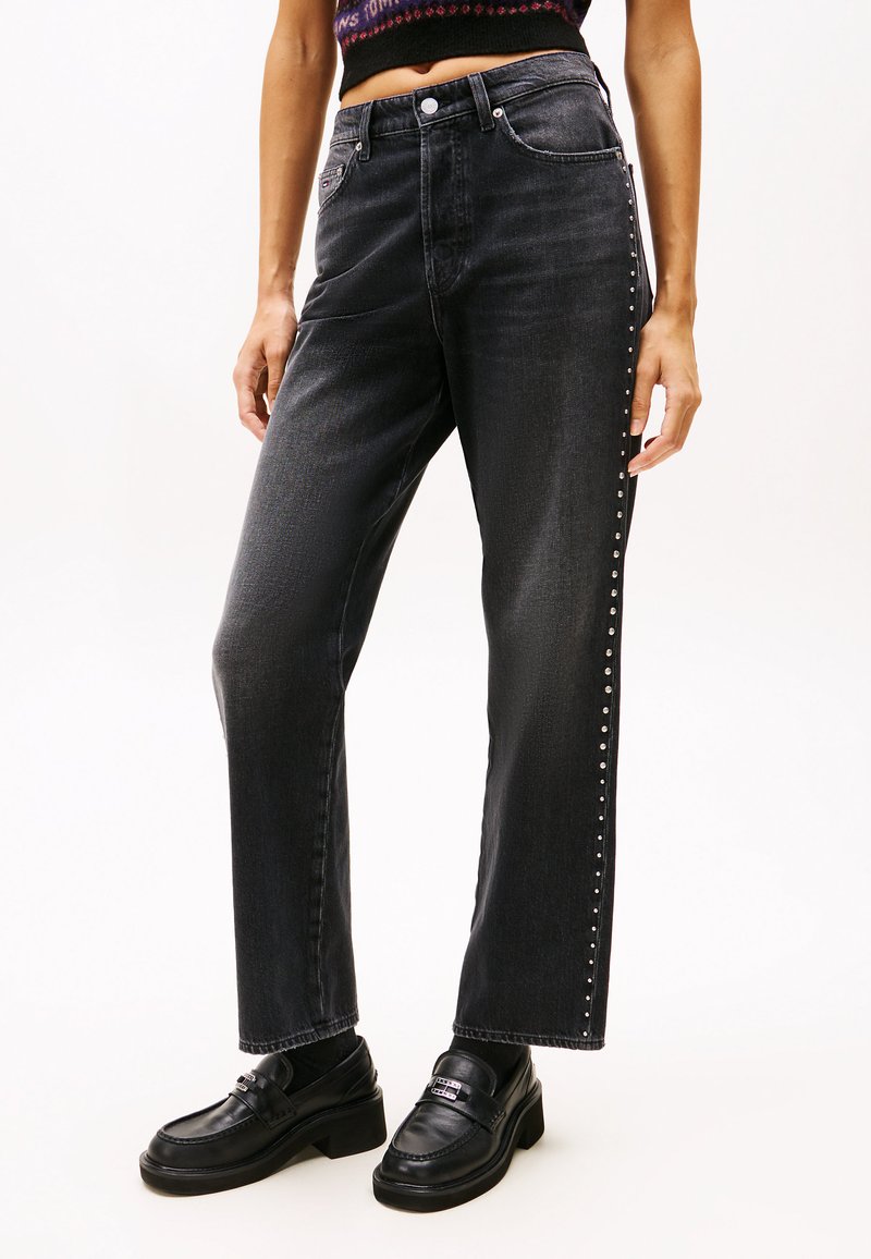 Jeans de mezclilla negros de tiro alto con corte recto, que presentan un detalle de tachuelas a lo largo del costado y una textura sutil. Combinados con mocasines negros.