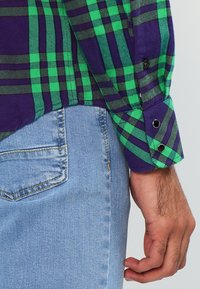 Zielono-fioletowa koszula w kratę z materiału flanelowego z czarnym guzikiem, zestawiona z jasnoniebieskimi dżinsami. Zbliżenie na rękę opartą na pasie.