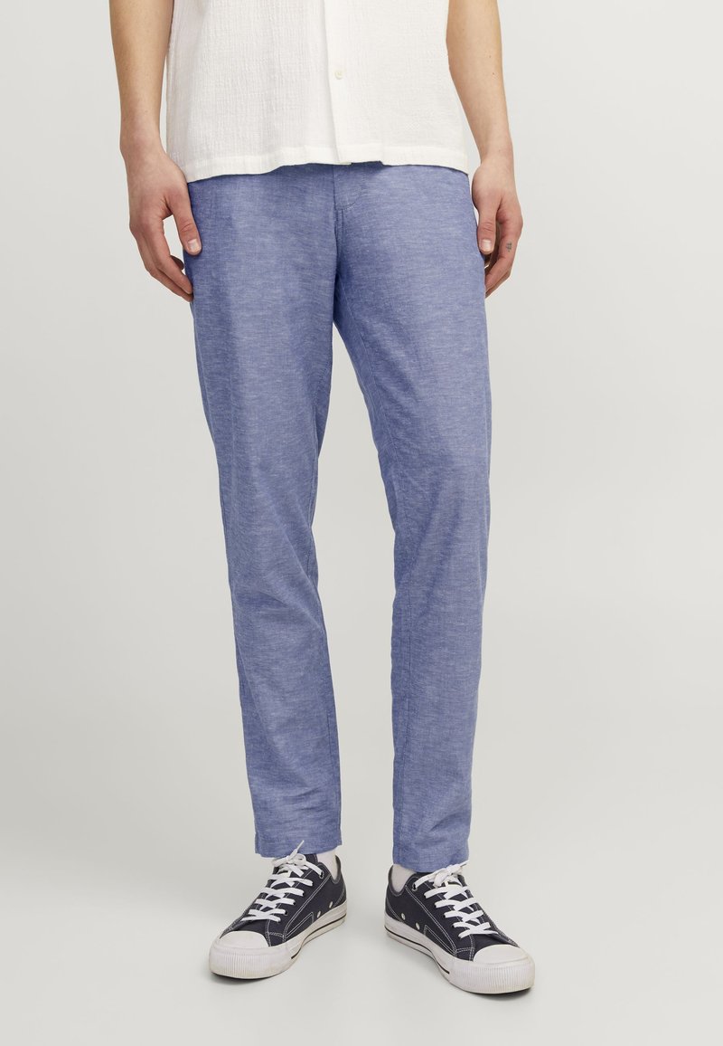 Pantalones de lino azul claro con una textura suave, corte ajustado y longitud hasta el tobillo. Combinados con zapatillas oscuras con suelas y cordones blancos.