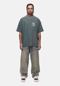 Przesadnie duża szara koszulka z prążkowaną teksturą i okrągłym grafiką na klatce piersiowej, zestawiona z jasnymi, luźnymi dżinsami i czarnymi sneakersami.