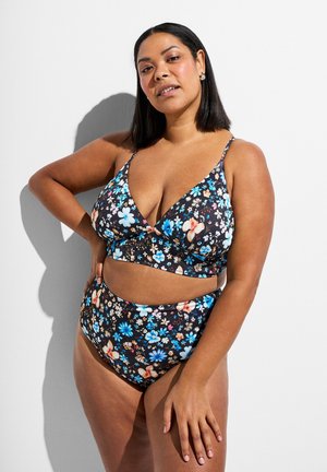 Woman with medium skin and straight black hair wearing a black floral print bikini, posing with hand on hip against a white background.