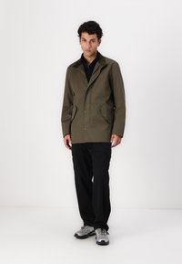 Olive green jacket with a snap button closure, two front pockets, and a collar design. Paired with black cargo pants and gray shoes.