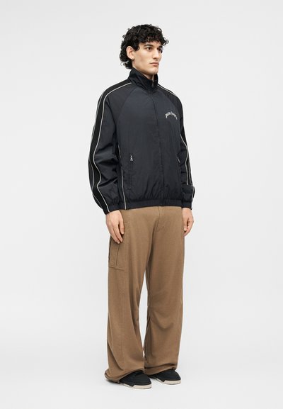 Noore mehe seljas on must lukuga jope valge servaga ja beežid laia säärega püksid, ta seisab ühevärvilise heledat tausta ees.