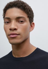 Close-up of a person with short, curly dark brown hair wearing a black crew-neck shirt. Smooth skin tone with neutral facial features.