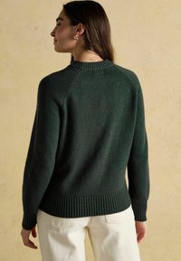 Dunkelgrüner gestrickter Pullover mit geripptem Saum und Bündchen, ausgestattet mit einem Rundhalsausschnitt und Raglanärmeln, vor einem hellbeigen Hintergrund.