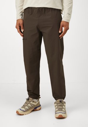 Osoba nosząca ciemnobrązowe spodnie dresowe z elastycznym pasem i kieszenią na zamek oraz beżowe buty trekkingowe na białym tle.