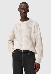 Krémový pletený sveter s textúrovaným dizajnom obsahujúcim káblové vzory a vertikálne pruhy. Nosí sa s čiernymi džínsami a opaskom.