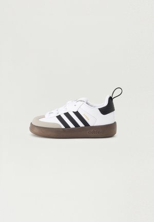 Biały sneaker Adidas Samba z czarnymi paskami, beżowym czubkiem, czarną zakładką na pięcie oraz brązową, teksturowaną podeszwą Adirom na jasnym tle.