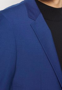 Blauwe blazer van een gestructureerde stof, met een notch-lapel en een slanke pasvorm, gecombineerd met een zwarte coltrui eronder.