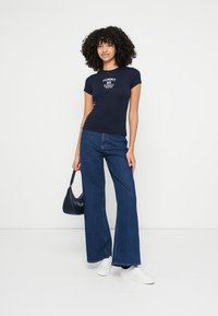 T-shirt azul-marinho justa com texto branco, combinada com jeans de perna larga. Modelo segura uma pequena bolsa preta e usa ténis brancos.