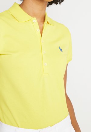 Femme portant un polo jaune vif à manches courtes avec un petit logo bleu brodé sur la poitrine gauche.