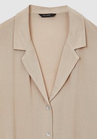 Camicia beige con colletto realizzata in un tessuto liscio e testurizzato. Presenta una chiusura con bottoni trasparenti, l'etichetta riporta la scritta "CALLIOPE," taglia S.
