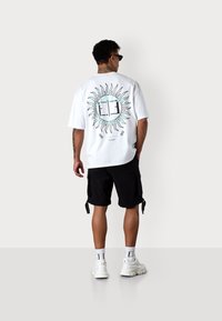 White T-shirt featuring a sun graphic in black with turquoise accents. Paired with black shorts and white sneakers. Clean, modern design.