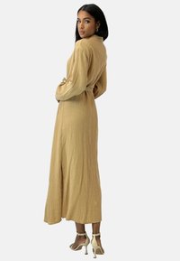 Długa beżowa sukienka z teksturowanej tkaniny, z wiązaniem w talii i bocznym rozcięciem. W połączeniu z jasnymi sandałami na obcasie.