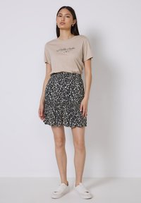 T-shirt beige avec impression noire, associé à une jupe à motifs noirs et crème avec des volants. Modèle portant des baskets blanches.