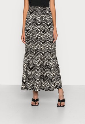Femme portant une longue jupe à motif chevron noir et blanc et des sandales noires à talons ouverts, debout devant un fond uni.