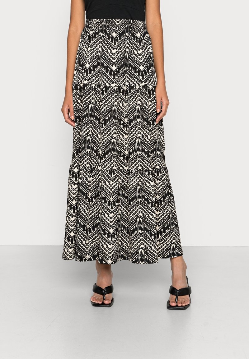 Femme portant une longue jupe à motif chevron noir et blanc et des sandales noires à talons ouverts, debout devant un fond uni.