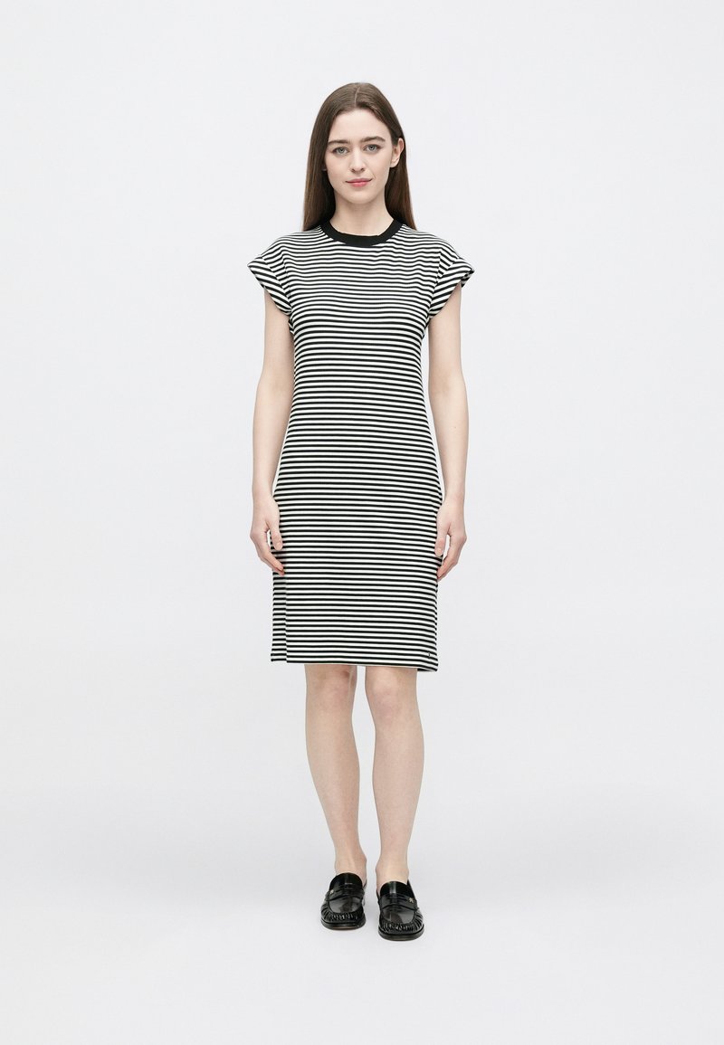 Zwart-wit gestreepte jurk met korte mouwen, tot op de knie, met een ronde halslijn. De stof oogt zacht en licht getailleerd. Zwarte platte schoenen.