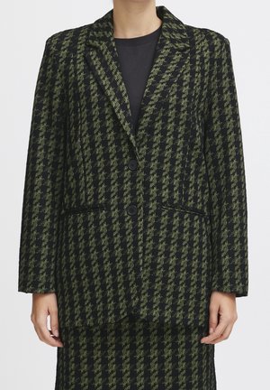 Kobieta ubrana w czarno-zieloną marynarkę w jodełkę z dwoma guzikami oraz dopasowaną spódnicę, pokazana od ramion do połowy uda.