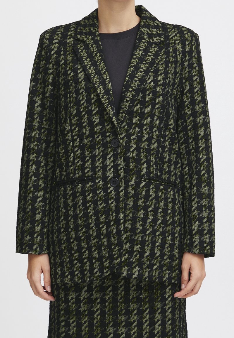 Femme portant un blazer pied-de-poule noir et vert à deux boutons avec une jupe assortie, vue des épaules jusqu'au milieu des cuisses.