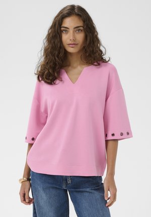 Kobieta o kręconych brązowych włosach, ubrana w luźny różowy top z dekoltem w kształcie litery V i oczkami na rękawach oraz niebieskie dżinsy, stojąca na białym tle.