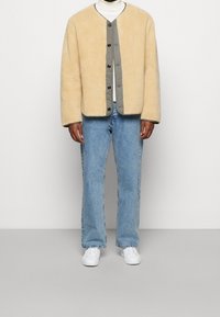 Giacca in pelliccia artificiale beige con foderatura grigia, bottoni sulla parte anteriore. Abbinata a jeans dritti azzurri e sneakers bianche.