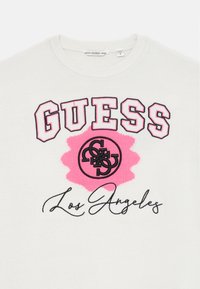 Vit stickad sweatshirt med fet rosa text "GUESS", en rosa grafisk detalj och svart text "Los Angeles".