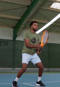 Tenis igralec v olivno zeleni majici s tiskom in svetlo modrih kratkih hlačah, drži oranžno teniško lopar, pripravljen na igro na igrišču.