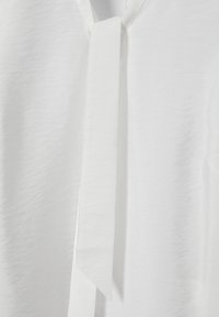 Gros plan sur un tissu blanc avec une bande verticale étroite de tissu suspendue, montrant une texture subtile et des détails de couture.