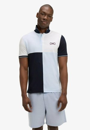 Homme portant un polo bicolore en bleu clair, blanc et bleu marine, assorti à un short bleu clair, debout devant un fond blanc.