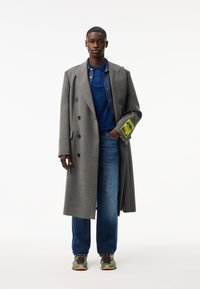 Cappotto lungo grigio in lana con design doppiopetto, grandi rever, abbinato a un polo blu, jeans in denim scuri e sneakers multicolori. Dettaglio del tag neon.