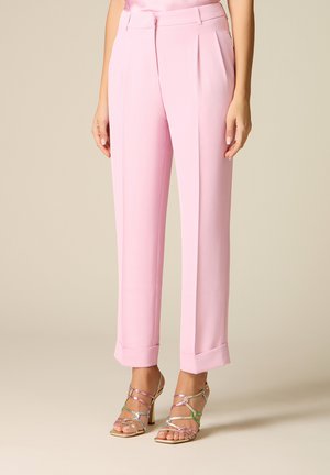 Kobieta ubrana w jasnoróżowe, wysokowadżone, dopasowane spodnie z mankietami oraz metaliczne, wielokolorowe sandały na wysokim obcasie z paskami.