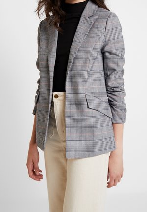 Mujer con un blazer de cuadros gris sobre un suéter de cuello alto negro y pantalones beige de talle alto, de pie frente a un fondo liso.