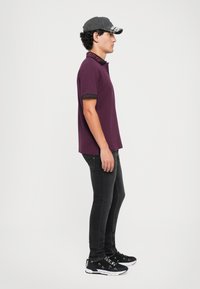 Violet polo marškinėliai su juodais akcentais, derinami su juodomis lieknomis džinsomis ir juodais sportbačiais, kuriuose yra baltas logotipas. Juos dėvi vyrų modelis.