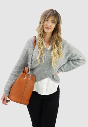 Vrouw met lang golvend haar, gekleed in een grijze gebreide trui over een wit blouse en zwarte broek, met een grote bruine leren handtas over haar schouder.