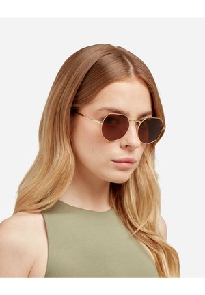 Gouden metalen zonnebril met ronde bruine glazen, voorzien van een dunne montuur. Model draagt een mouwloos olijfgroen topje en heeft lang blond haar.