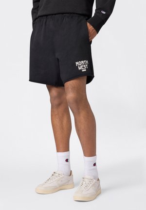 Hombre con pantalones cortos negros con texto "NORTH WEST", calcetines deportivos blancos con logo y zapatillas de color blanco roto, de pie frente a un fondo liso.
