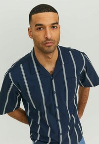 Tamsiai mėlyna trumpų rankovių marškiniai su baltais vertikaliais dryžiais, sagčių priekyje, įpjovimu ant apykaklės ir vienu krūtiniu kišenėje. Lengvos medžiagos tekstūra.