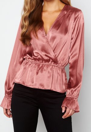 Femme portant un chemisier en satin rose à manches longues, avec un col en V croisé et une taille froncée, associé à un pantalon noir.