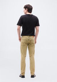 Νέος άντρας που στέκεται με την πλάτη, φοράει μαύρο μπλουζάκι, μπεζ τζιν και μαύρα παπούτσια μπροστά σε απλό λευκό φόντο.