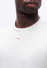 Κοντινή λήψη ενός ατόμου που φοράει λευκό μπλουζάκι με στρογγυλή λαιμόκοψη και μικρό κόκκινο και λευκό λογότυπο Reebok στο στήθος.