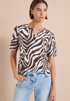 Frau, die ein kurzärmeliges, zebragestreiftes Oberteil und hellblaue Jeans trägt und mit einer Hand in der Tasche vor einem beigen Hintergrund steht.