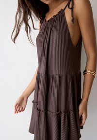 Brown ribbed dress with a gathered neckline, adjustable straps, and a ruffled hem. Features a relaxed fit and textured fabric.