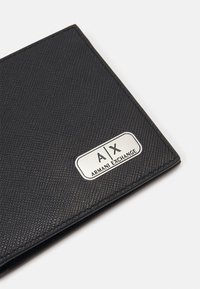 Crna teksturirana novčanica napravljena od sintetičkog materijala sadrži srebrnu pločicu s logom 'A/X ARMANI EXCHANGE' i geometrijski uzorak.