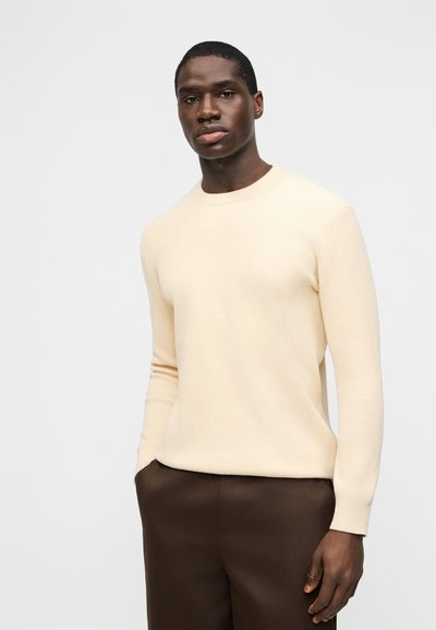 Homme portant un pull crème à manches longues et un pantalon marron foncé, debout les mains dans les poches sur un fond clair uni.