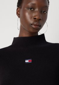 Svart ribbestrikket genser med høy hals; har et lite rødt, hvitt og blått logo-patch på venstre side. Glatt tekstur, moderne design.